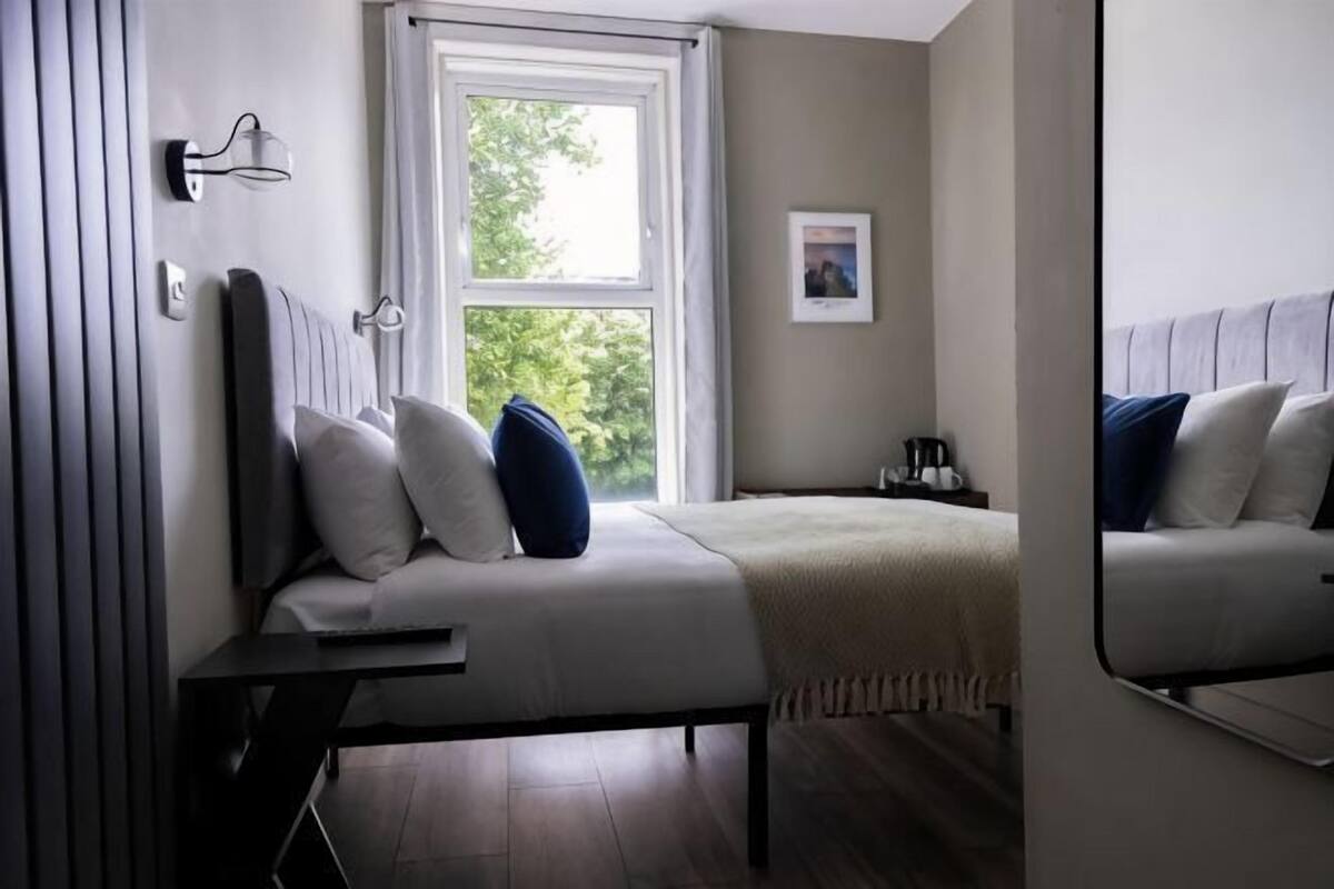 A bedroom with a bed, bedside table, lamp, window, and a framed picture on the wall.