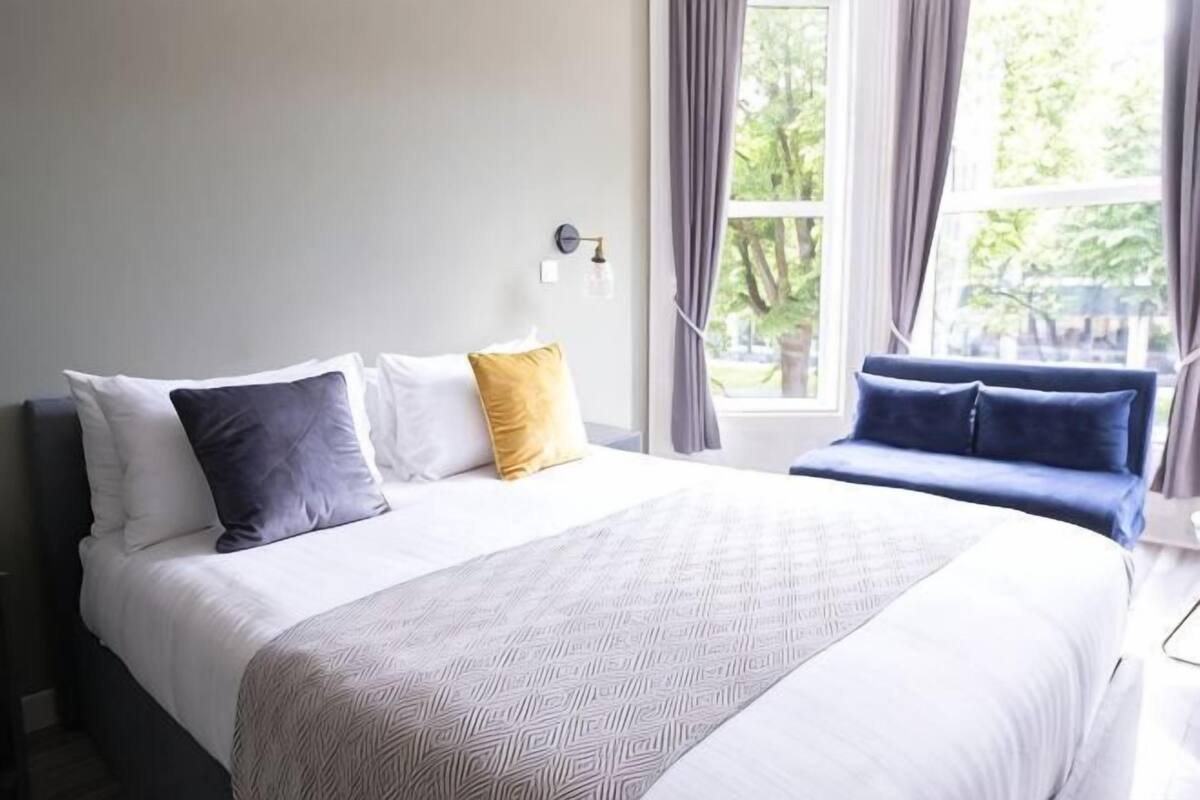 A neatly made bed with pillows, a bedside table, and a window with curtains.