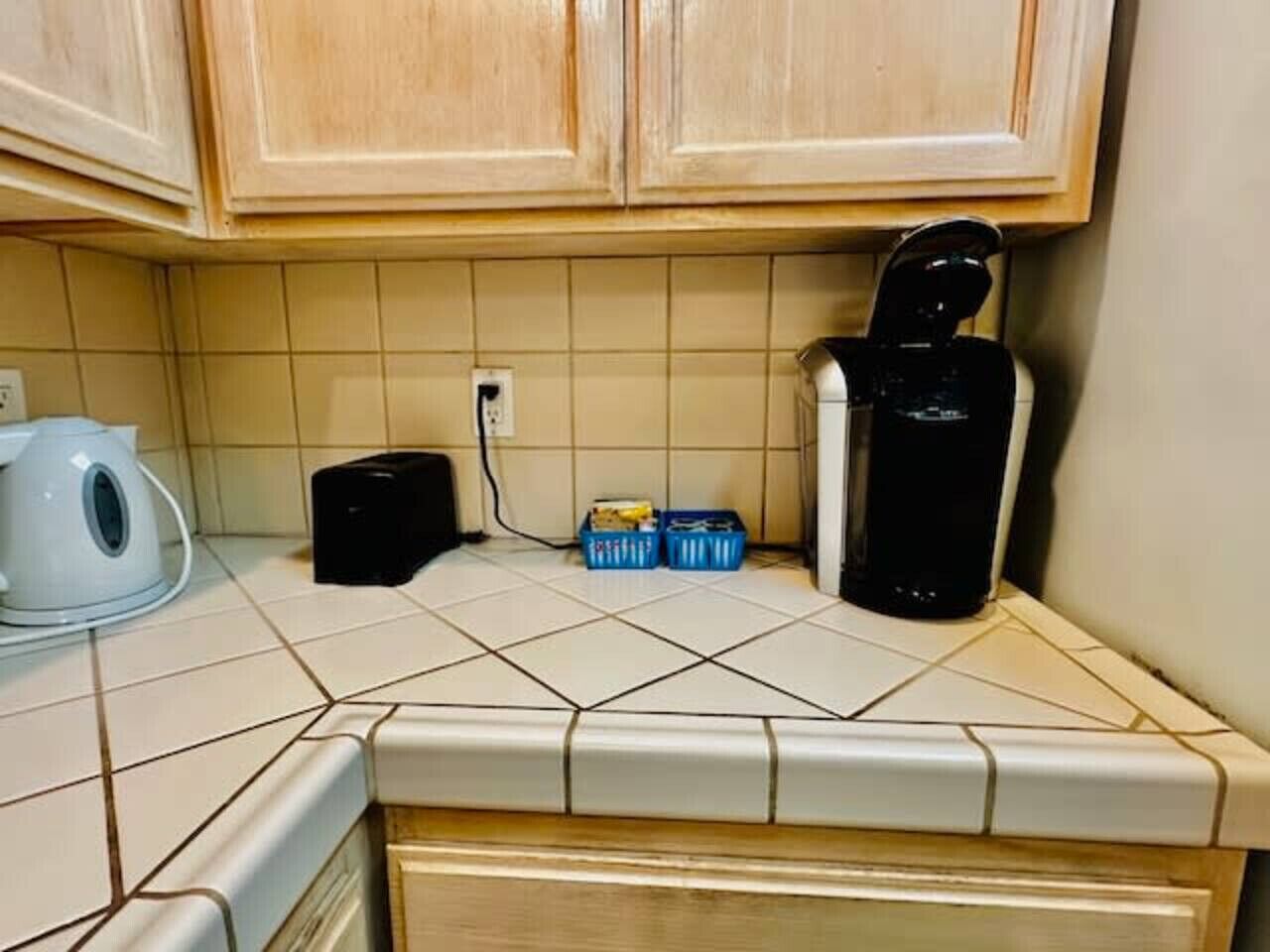 A kitchen with white tiles, wooden cabinets, a kettle, a coffee maker, and two blue containers.