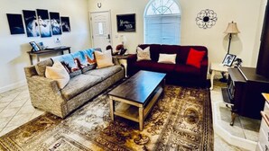 A living room with a sofa, a coffee table, and a red armchair. There is a window with blinds, a ceiling fan, and a rug on the floor.