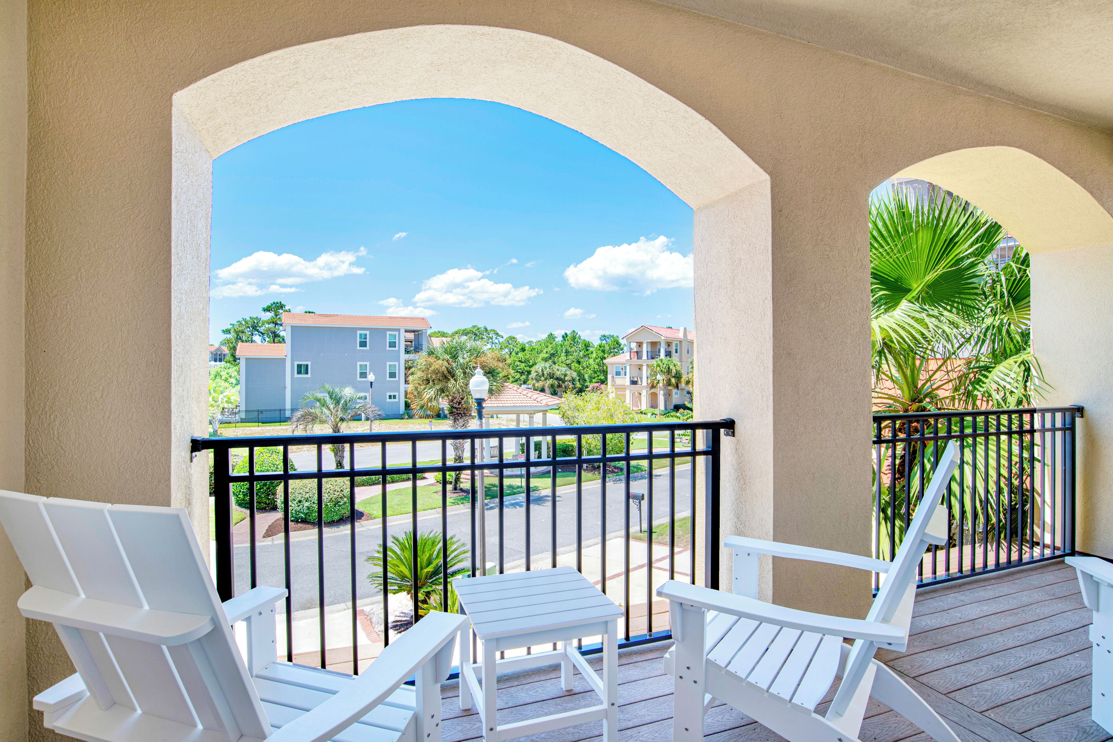 A balcony with white chairs and a table, overlooking a residential area with houses and palm trees.