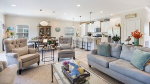A spacious living room with a grey sofa, armchairs, and a coffee table. There is a dining area with a table and chairs, and a kitchen area with white cabinets and appliances.