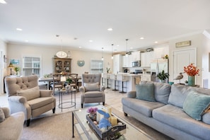 A spacious living room with a grey sofa, armchairs, and a coffee table. There is a dining area with a table and chairs, and a kitchen area with white cabinets and appliances.