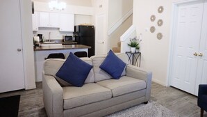 Un salon avec un canapé beige, deux coussins bleus, une petite table d’appoint avec une plante, et un escalier menant à un niveau supérieur.