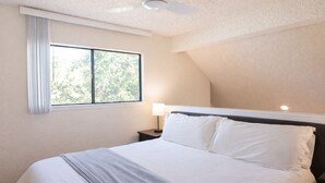 Une chambre à coucher comprenant un lit, une table de chevet, une lampe, une fenêtre avec des rideaux et un ventilateur de plafond.