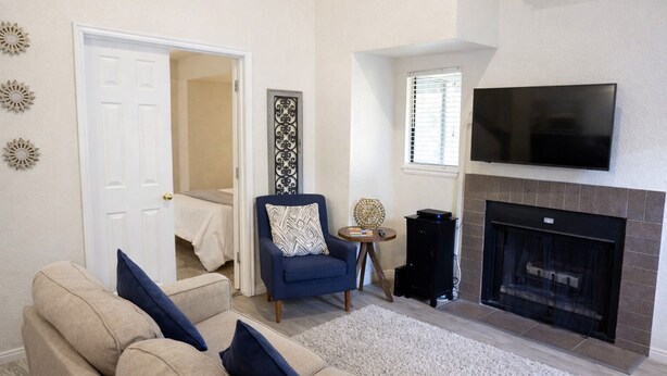 Un salon avec un canapé beige, un fauteuil bleu, une cheminée, un téléviseur fixé au-dessus de la cheminée et une porte donnant sur une chambre.