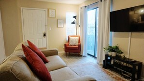A living room with a beige sofa, red cushions, a red armchair, a black TV stand with a plant, and a door with a white frame. - Down Town Convenience, TRAX, Self Check-In Studio (Salt Lake City)