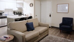 A living room with a beige sofa, a small round table, a dark armchair, a kitchen in the background, and a white door. - King Bed*Extensive Cleaning Protocol*Self Check-In (Salt Lake City)