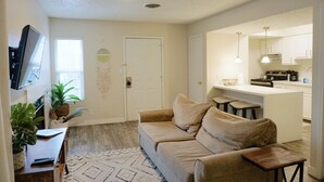 Sala de estar con sofá, mesa de centro y televisor. Zona de cocina con estufa y lavaplatos.