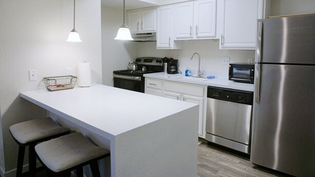 Una cocina moderna con armarios blancos, electrodomésticos de acero inoxidable, una isla central con taburetes y luces colgantes, además de un lavavajillas.