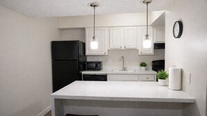 Cocina compacta con armarios blancos, refrigerador negro, microondas, pileta y dos taburetes.