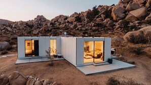 Two modern, light-colored cabins with glass doors, set in a rocky, arid landscape.