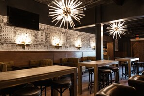 Interno di un ristorante con tavoli alti, banconi e sgabelli da bar. Sono presenti luci a parete e un grande murale raffigurante una veduta cittadina.
