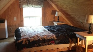 A cozy attic bedroom with a sloped ceiling, a bed with a patterned blanket, a bedside table with a lamp, and a window with a patterned curtain. - Micro house cabin located near Silver Mines Recreation Area. (Fredericktown)