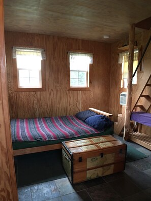Een klein houten huisje met een enkel bed, twee ramen, een houten kist en een trap.