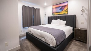 Ein modernes Schlafzimmer mit einem großen Bett, einem dunklen Kopfteil und einem Gemälde, das einen Sonnenuntergang über einer Klippe zeigt.