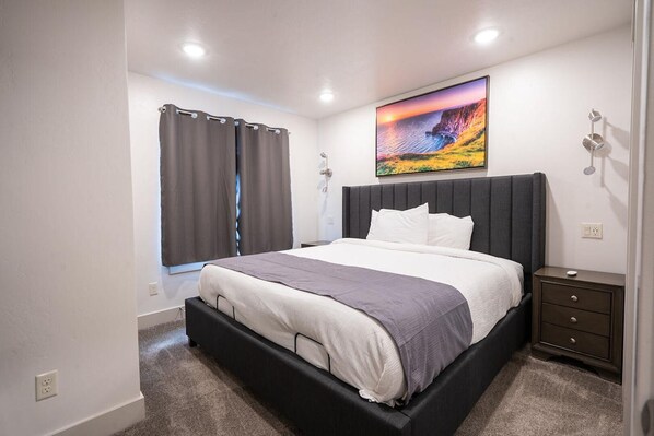 A modern bedroom with a large bed, dark headboard, and a painting of a sunset over a cliff.