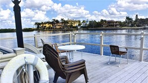 Une terrasse avec une table et deux chaises, une bouée de sauvetage et un garde-corps donnant sur une étendue d’eau, avec des bâtiments en arrière-plan.