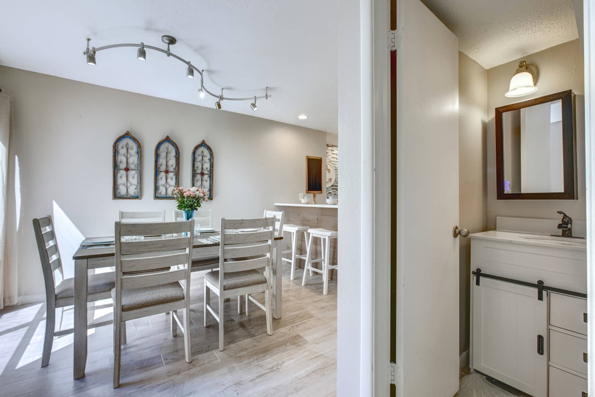 A dining area with a wooden table and chairs, a vase with flowers, and a wall-mounted light fixture.