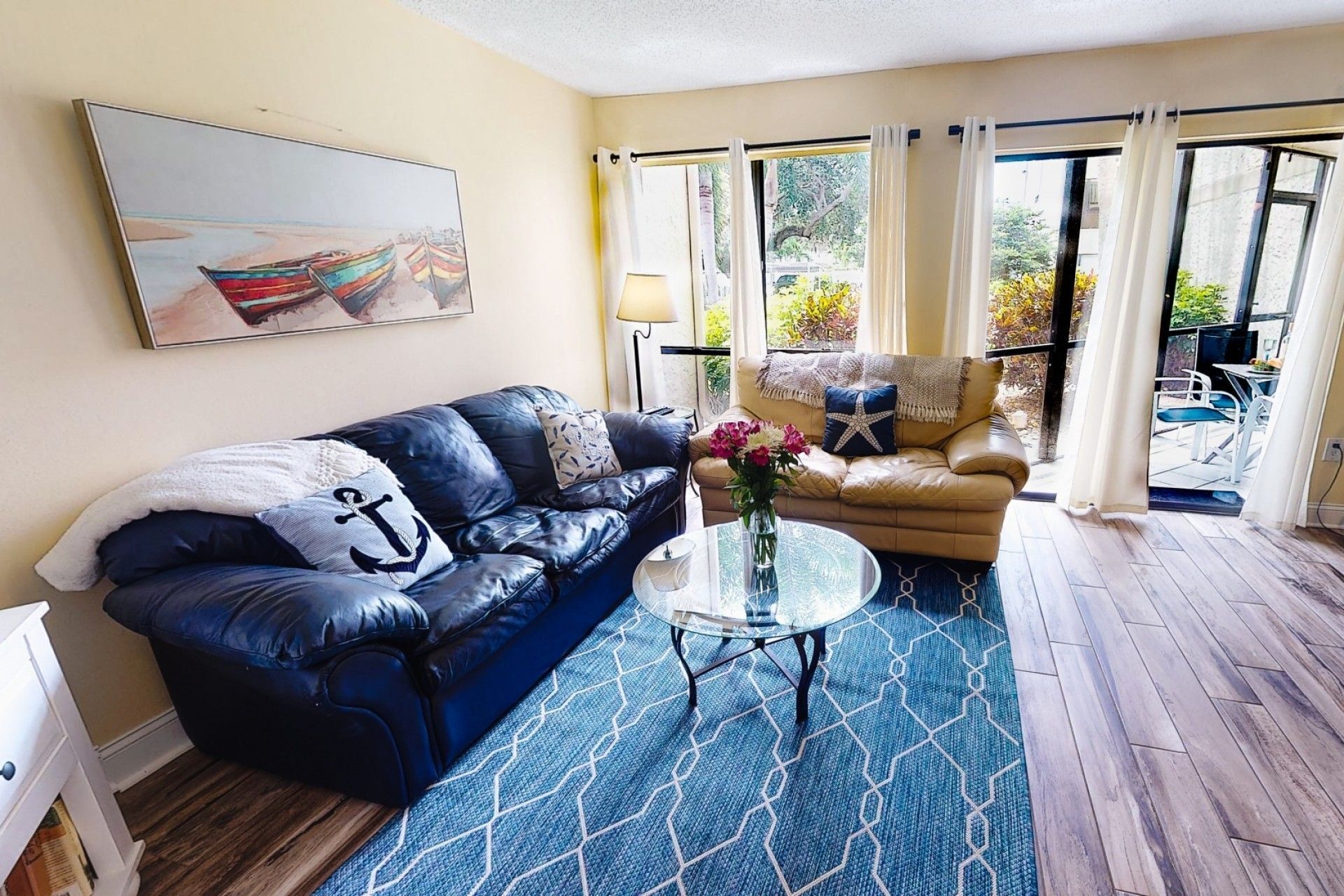 A living room with a blue leather sofa, a glass coffee table, and a beige sofa. There is a painting on the wall and a window with curtains.
