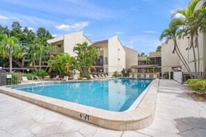 A swimming pool with lounge chairs, palm trees, and a multi-story building with balconies. - Bay Oaks C Ground Floor (Siesta Key)