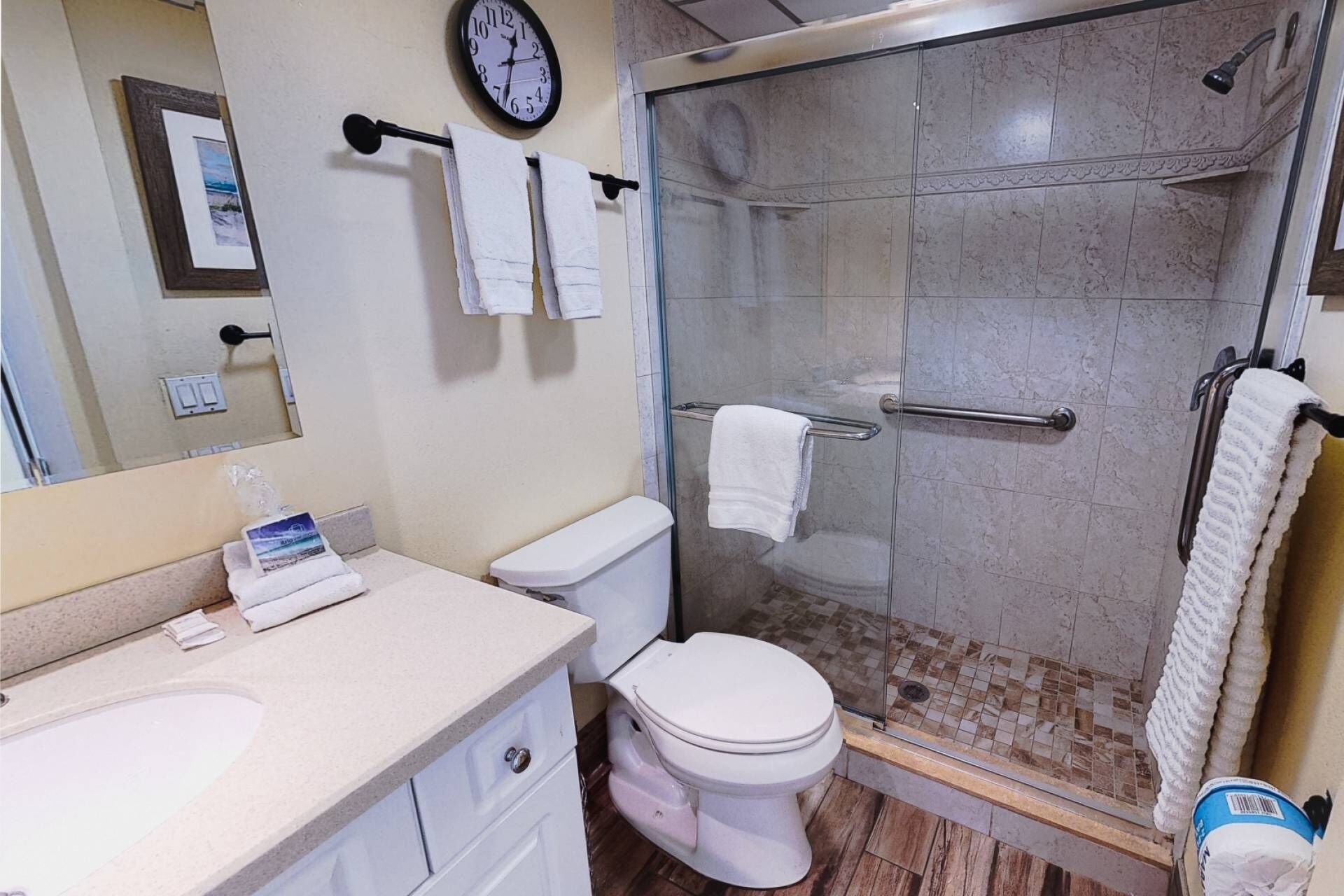 Une salle de bain avec des toilettes, un lavabo et une douche. Il y a une horloge au mur et des serviettes suspendues sur un porte-serviettes.