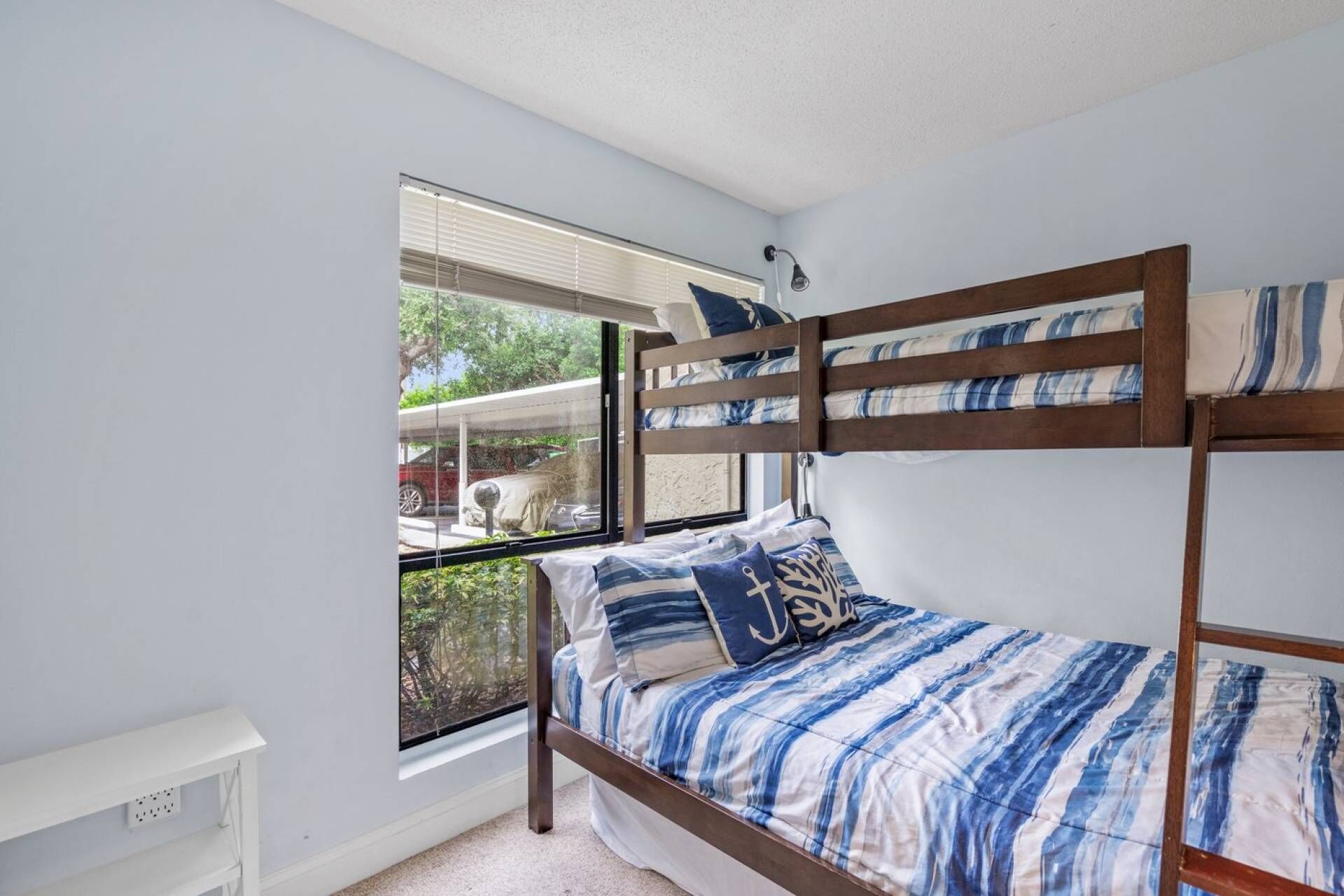 Una cama litera con ropa de cama azul y blanca a rayas, una ventana que ofrece vistas a la vegetación y un coche aparcado afuera, y una pared blanca con toma de corriente.