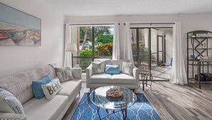 A modern living room with a sofa, a glass coffee table, and a view through large glass doors to an outdoor area. - Bay Oaks C Ground Floor (Siesta Key)