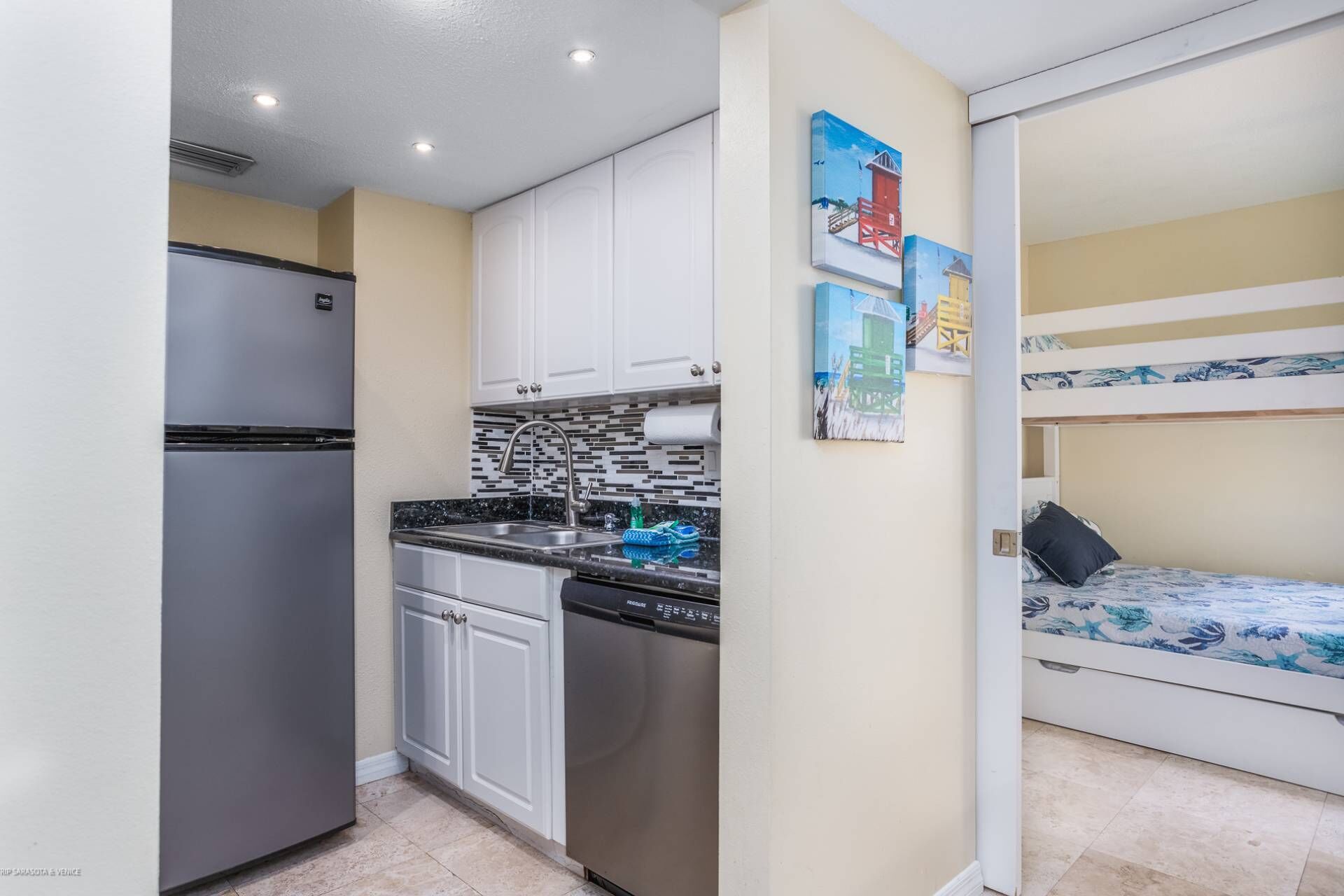 A compact kitchen with white cabinets, a stainless steel refrigerator, and a tiled backsplash. Adjacent to the kitchen is a bunk bed with a floral bedspread.