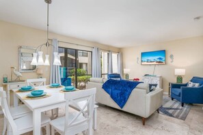 A modern living room with a dining area, featuring a sofa, a TV, and a dining table with blue plates. - Bay Oaks A Ground Floor (Siesta Key)