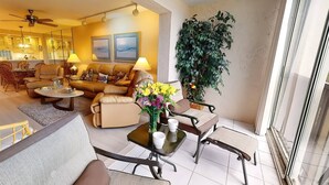 A spacious living area with a sofa, armchairs, a coffee table, and a dining table. There is a large potted plant and a view of the kitchen in the background. - Bay Oaks, 4th Floor Corner - Elevator (Siesta Key)