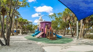 Un parque infantil con toboganes y estructuras para escalar, rodeado de palmeras y un cielo despejado.