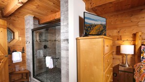 Interno di una baita in legno con doccia in vetro, armadietti in legno e un letto con coperta colorata.