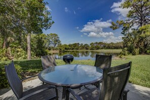 A round glass table with four chairs, a black grill, and a view of a lake surrounded by trees.