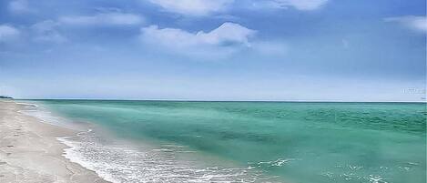 A beach with clear turquoise water and a sandy shore under a blue sky with clouds.