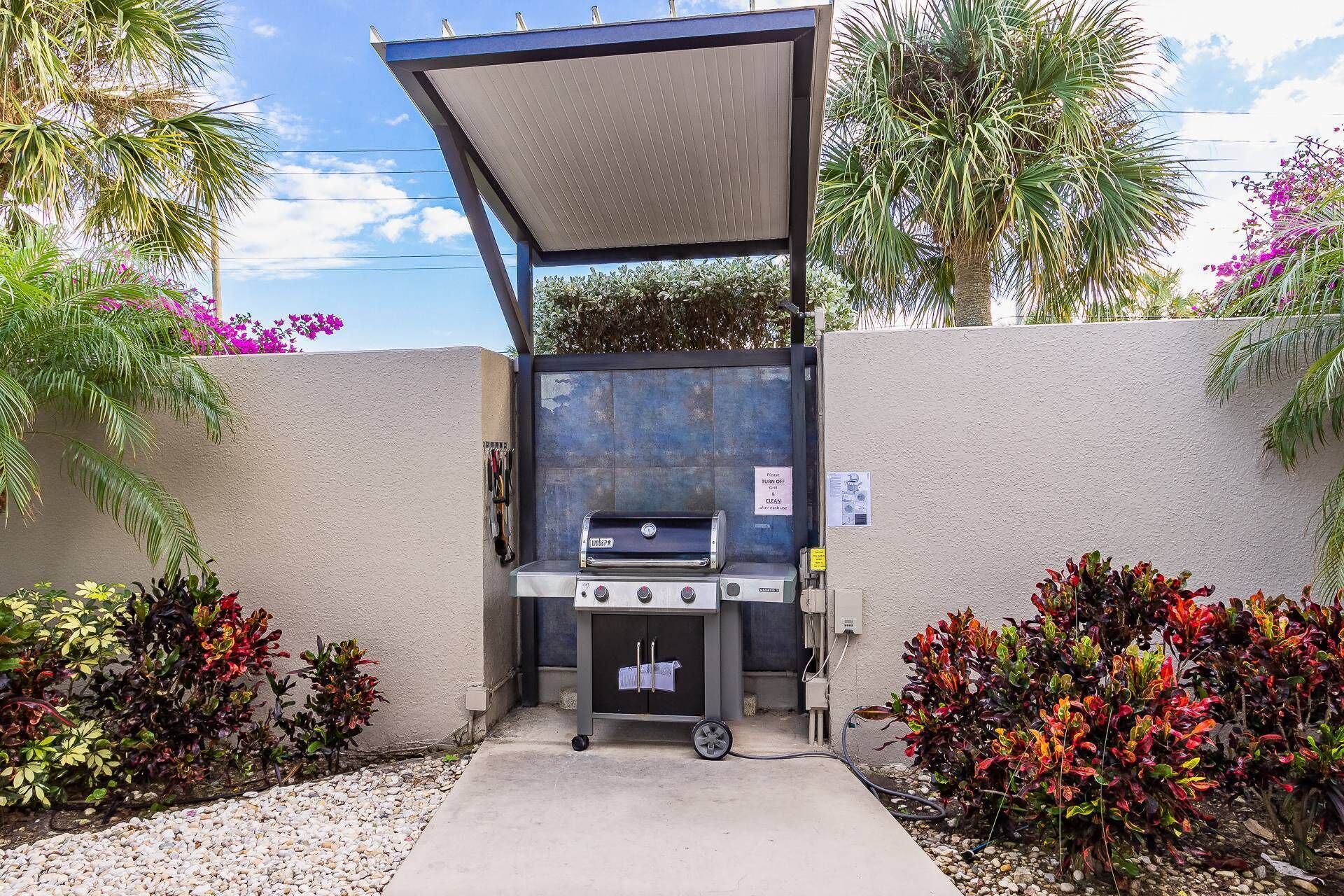 An outdoor grill area with a canopy, surrounded by decorative plants and a concrete pathway.