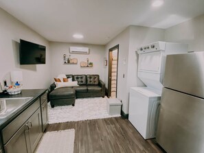 A compact kitchen with a built-in oven, a refrigerator, and a sink. There is a dark-colored sofa with cushions and a white rug in the center.