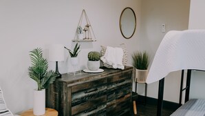 A bedroom with a wooden dresser, a bedside table with a lamp, a hanging plant, and a bed with a white comforter. - City Loft  (Trenton)