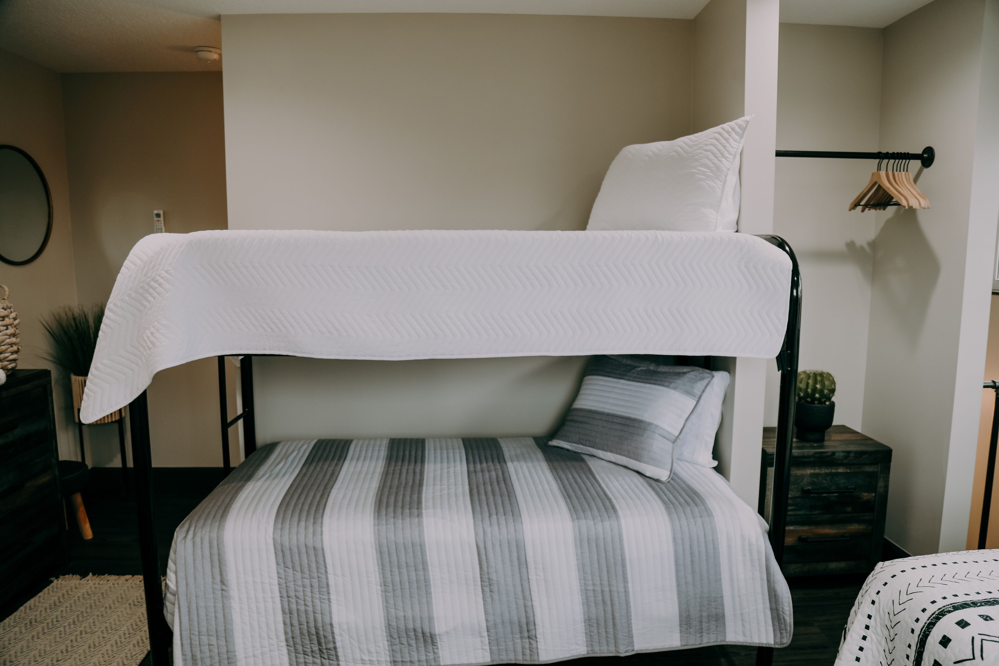 Un lit superposé avec un lit supérieur blanc et un lit inférieur à rayures. Il y a une petite table d’appoint avec une plante et un porte-vêtements en métal noir sur lequel sont suspendus des vêtements.