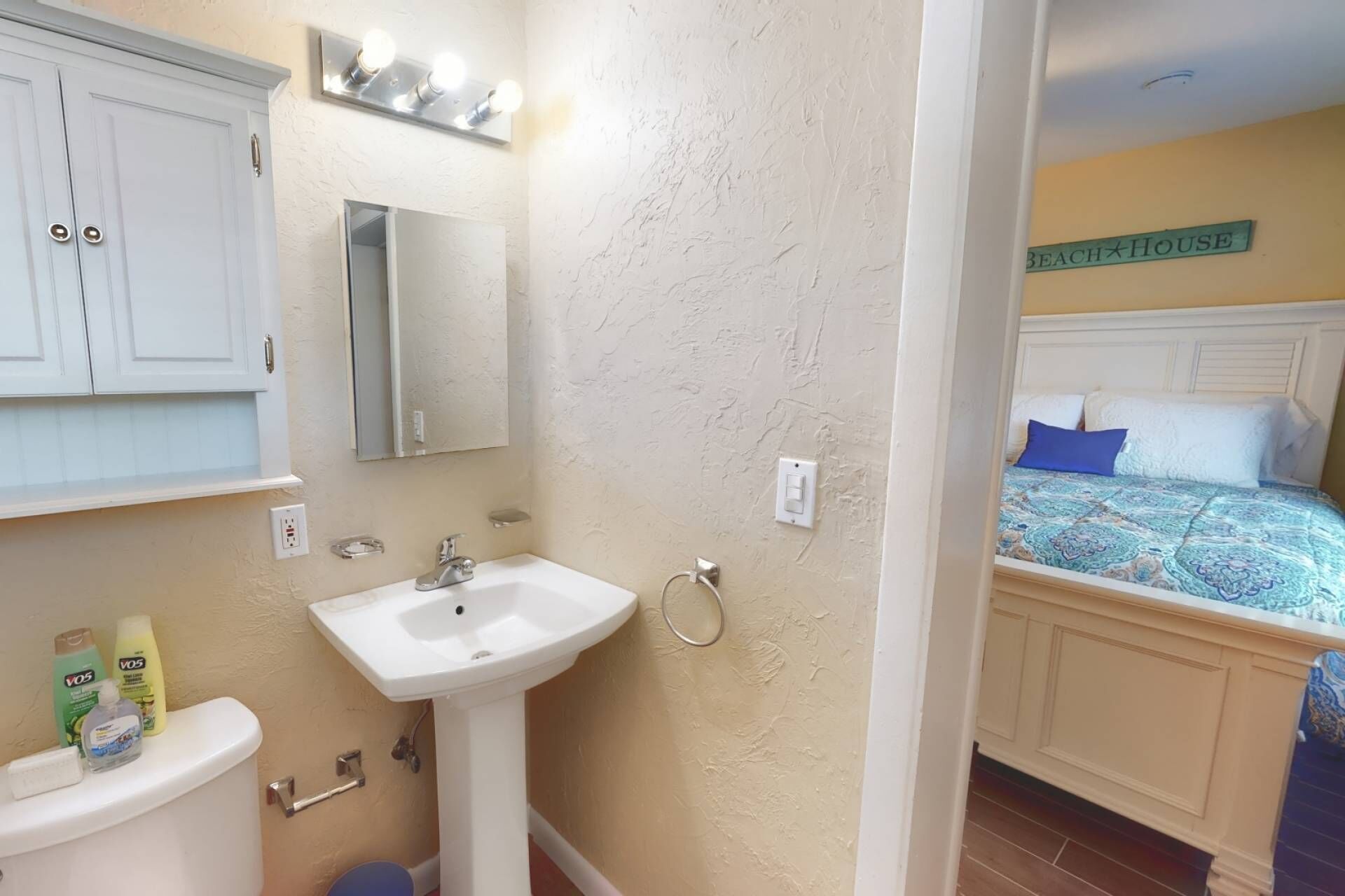 A bathroom with a sink, mirror, and toiletries. Adjacent is a bedroom with a bed and a sign reading "Beach House".