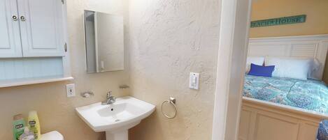A bathroom with a sink, mirror, and toiletries. Adjacent is a bedroom with a bed and a sign reading "Beach House".