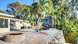 A pool area with a brick fire pit, palm trees, and a building with a blue roof. - Peacock Bungalow - Left (Siesta Key)