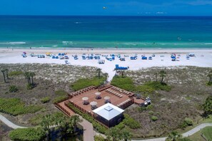 An aerial view of a beach resort with a large sandy beach, numerous umbrellas, and a central building with outdoor seating and a pool. - Siesta Dunes on Siesta Key (Siesta Key)