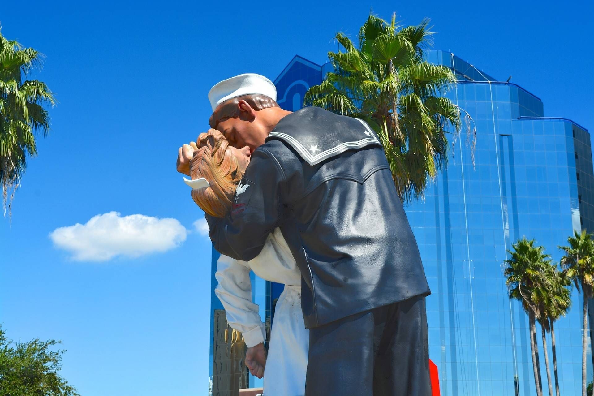 Une statue représentant deux personnages qui s’enlacent, avec un bâtiment moderne et des palmiers en arrière-plan.