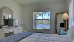 Un dormitorio con una cama, un televisor sobre una cómoda, una ventana con vista a un lago y una lámpara en una mesita de noche.