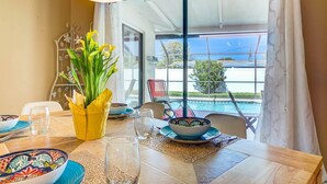 Ein Essbereich mit einem Tisch für zwei Personen, einer Vase mit gelben Blumen und Blick auf einen Swimmingpool und Grünflächen außerhalb.