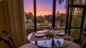 A dining table set for two with a view of a sunset over a lake, surrounded by chairs and a glass table. - White Sands Walk to Siesta Beach (Siesta Key)