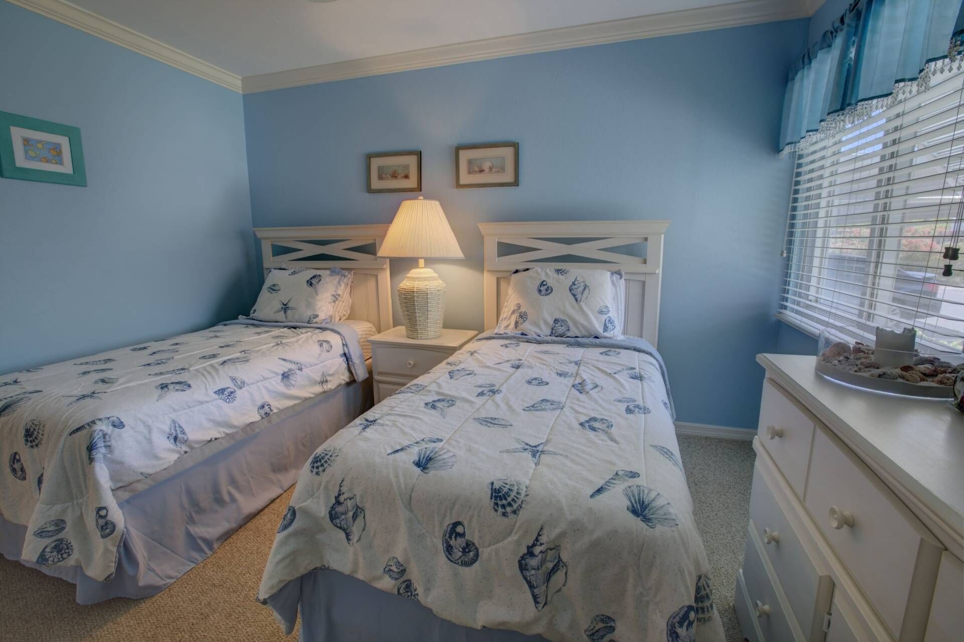 A bedroom with two beds, a nightstand with a lamp, a dresser, and a window with blinds.