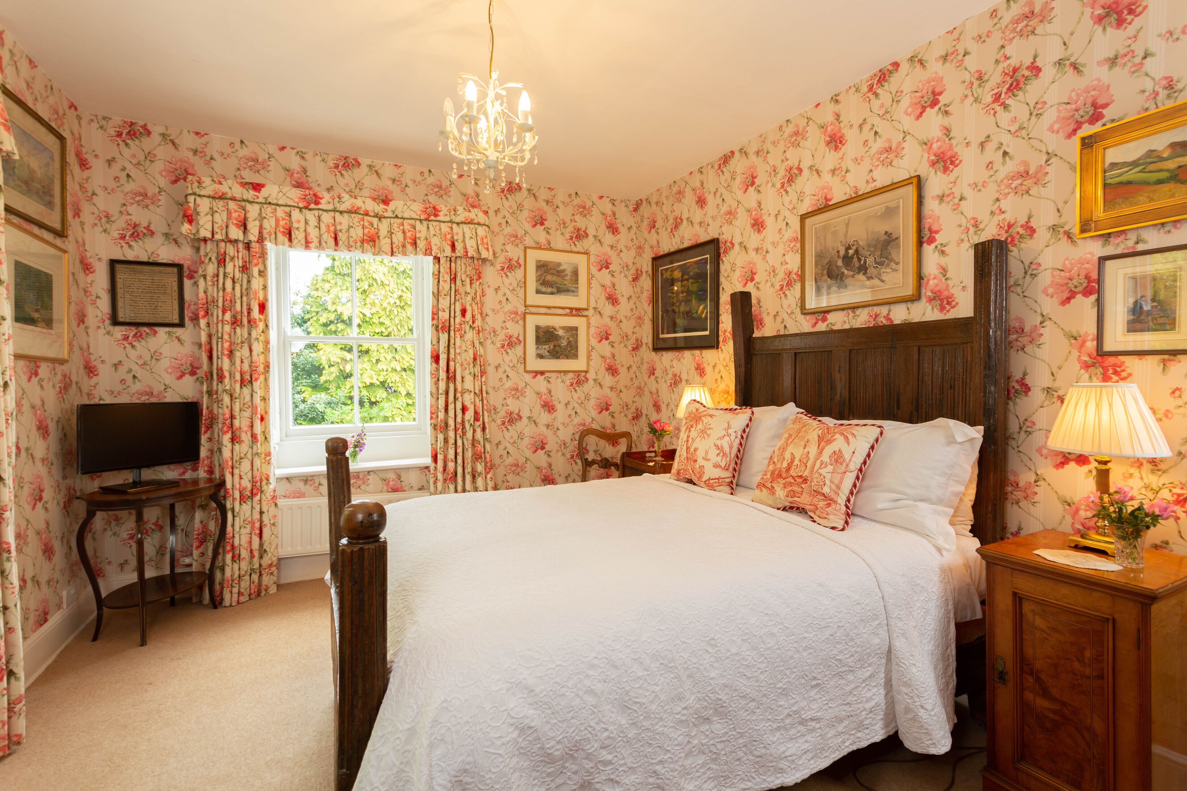 A bedroom with floral wallpaper, a wooden bed, a television on a side table, and a window with curtains.