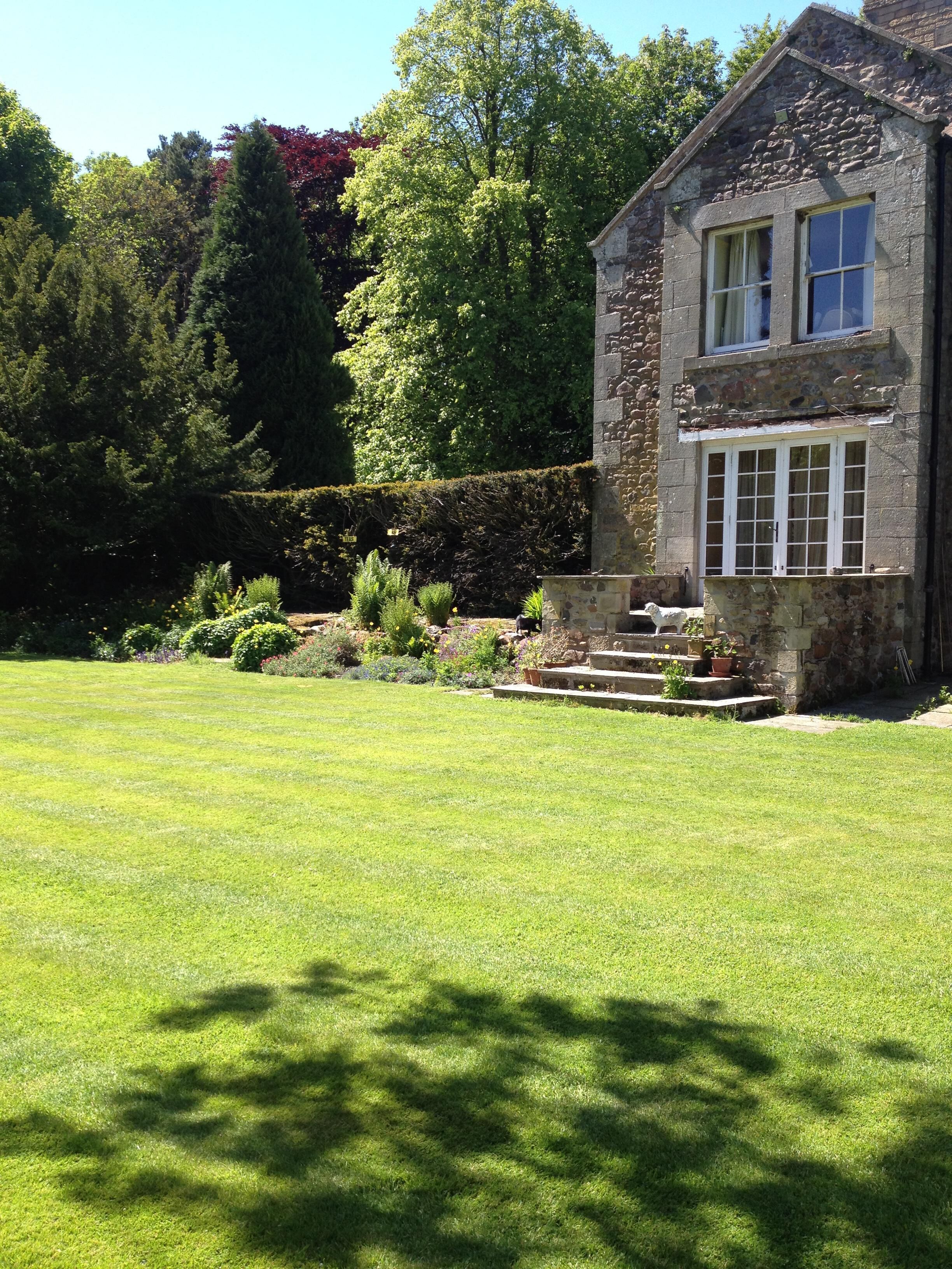 A stone building with a well-maintained lawn, steps leading to the entrance, and a hedge in the background.